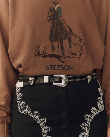 A person wearing a brown Stetson sweatshirt with a cowboy graphic, THE GREAT. F26 STETSON “The Western Hardware Belt” in black with a silver buckle, and a black skirt featuring white embroidered patterns.