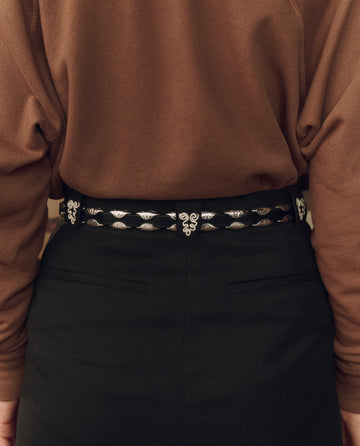 Person wearing a brown long-sleeve top, black high-waisted pants, and THE GREAT. F26 STETSON The Western Hardware Belt in Black with Silver, featuring silver decorative details and a silver-tone buckle, shown from the back.