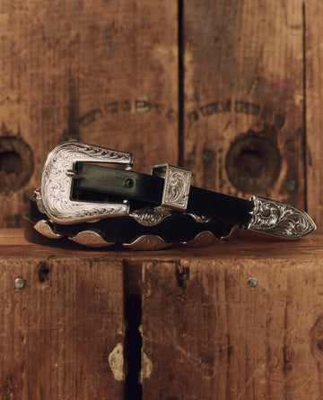 The Western Hardware Belt in black top-grain leather from THE GREAT. F26 STETSON features an ornate silver-tone buckle, tip, and loop, and is displayed coiled on a rustic wooden surface with visible grain and holes—a classic western accessory.