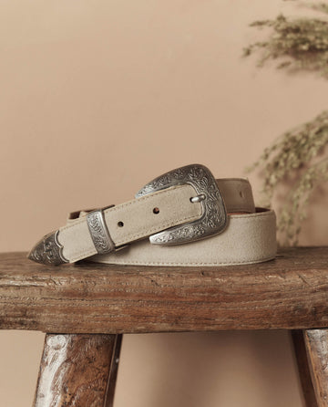 The Western Narrow Belt by THE GREAT. STETSON SP26, in cream with a silver engraved buckle, tip, and loop, is coiled on a rustic wooden stool against a neutral backdrop with dried foliage.