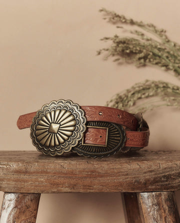 The Tooled Leather Concho Belt in Cognac With Brass by THE GREAT. STETSON SP26 is displayed on a wooden stool, featuring embossed patterns and ornate brass buckles, with dried foliage in the background.