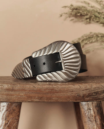 The Big Buckle Belt in black with a large silver buckle by THE GREAT. STETSON SP26 rests on a rustic wooden stool, with dried foliage in the background.