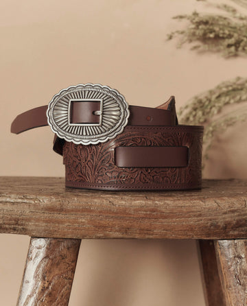 The Tooled Waist Belt in brown with silver by THE GREAT. STETSON SP26 features embossed floral patterns and an ornate buckle, displayed on a rustic wooden stool against a beige background.