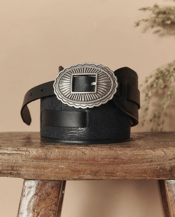 The Tooled Waist Belt in black with a silver buckle by THE GREAT. STETSON SP26 is coiled on a wooden stool and displayed against a neutral background.