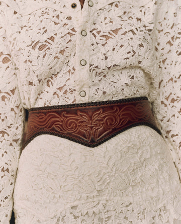 Close-up of a person wearing a white lace buttoned top and skirt, accessorized at the waist with THE GREAT. F26 STETSON’s The Tooled Corset Belt in Burnished Brown.