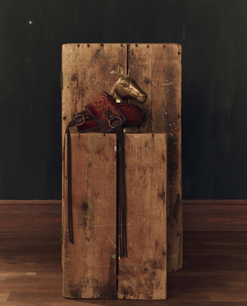 A burnished brown Tooled Corset Belt by THE GREAT. F26 STETSON rests atop a tall, weathered wooden box against a dark wall and wooden floor.
