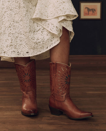 Wearing THE GREAT. F26 STETSON’s “The Southwest Cowboy Boot” in Chestnut with Turquoise Sunburst, a person stands on a wooden floor in a white lace skirt, with a framed horse picture adorning the wall in the background.