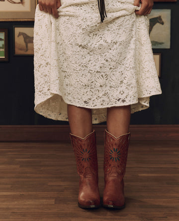 A person in a white lace dress and THE GREAT. F26 STETSON’s The Southwest Cowboy Boot in Chestnut with Turquoise Sunburst stands on a wooden floor, with framed horse pictures on the wall behind them.