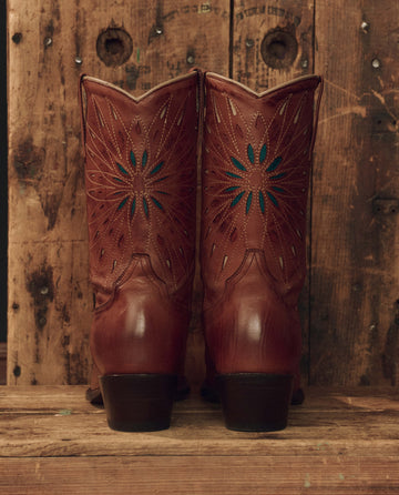 A pair of The Southwest Cowboy Boots by THE GREAT. F26 STETSON, crafted in rich chestnut leather with turquoise sunburst detailing, stand on a wooden surface against a rustic wooden background.