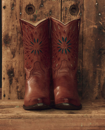 A pair of “The Southwest Cowboy Boot – Chestnut with Turquoise Sunburst” by THE GREAT. F26 STETSON, featuring handcrafted rich brown leather and decorative stitching, stands on a rustic wooden surface against a weathered wood backdrop.