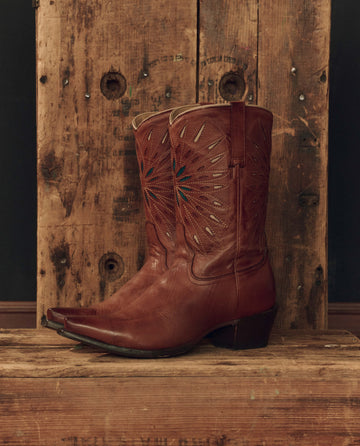 A pair of The Southwest Cowboy Boot -- Chestnut with Turquoise Sunburst by THE GREAT. F26 STETSON, handcrafted from rich brown leather with striking turquoise sunburst accents and decorative stitching, sits on a rustic wooden surface against a wood backdrop.