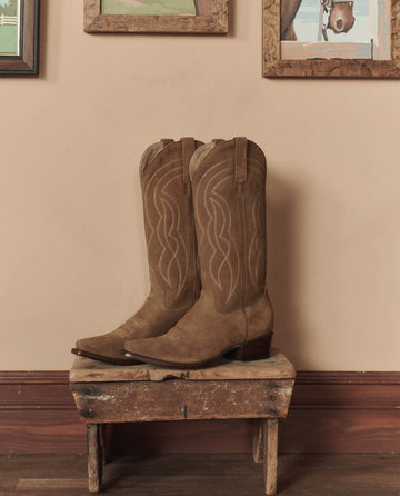 The Cowboy Boot (Suede) in tan by THE GREAT. STETSON SP26 sits on a wooden stool with a beige wall and framed art, showcasing classic western style with stitched patterns.