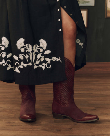 A person wearing THE GREAT. F26 STETSON’s The Basket Weave Cowboy Boot in oxblood and a black skirt with white floral embroidery stands on a wooden floor.