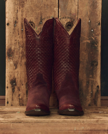 A pair of The Basket Weave Cowboy Boots in Oxblood by THE GREAT. F26 STETSON stands on a rustic wooden surface against a matching wooden background.