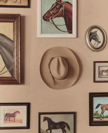 The Gus 6X Hat in Butterscotch by THE GREAT. STETSON SP26 hangs on a beige wall, surrounded by framed paintings and horse illustrations.
