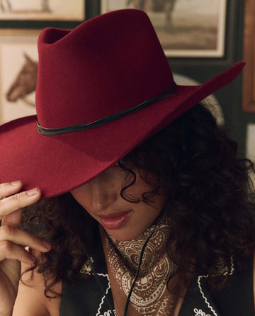 A person with curly hair wears THE GREAT. F26 STETSON The Ridge Hat—Maroon (limited edition) and a patterned bandana, tilting the hat downward. Framed pictures of horses can be seen in the background.