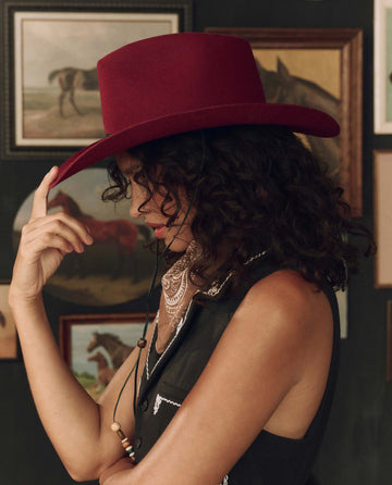 A person with curly hair, dressed in a sleeveless shirt, stands sideways touching the brim of The Ridge Hat in maroon by THE GREAT. F26 STETSON. Horse paintings can be seen in the background.