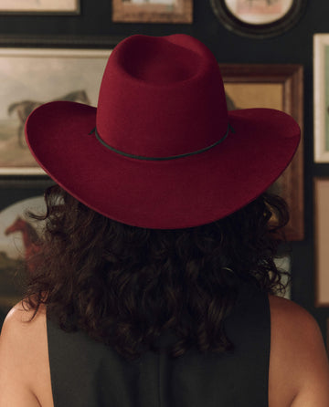 A person with curly dark hair, shown from behind and wearing a black top, models THE GREAT. F26 STETSON’s limited-edition The Ridge Hat in Maroon; framed pictures decorate the wall in the background.