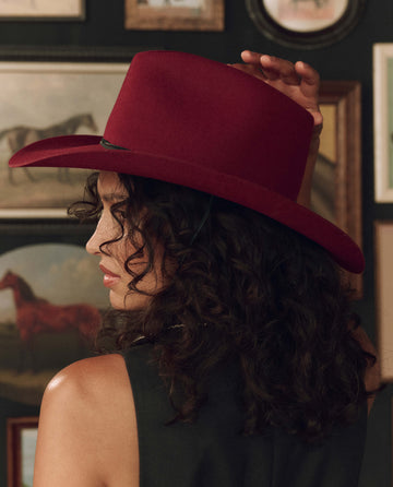 A person with curly hair, seen in profile against a wall adorned with framed art—including a painting of a horse—is wearing THE GREAT. F26 STETSON “The Ridge Hat” in Maroon.