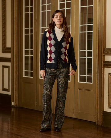 A person wears The Argyle Menswear Cardigan. -- Navy in a cream, maroon, and grey argyle pattern over a cream turtleneck, paired with patterned trousers.