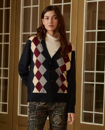 A person wearing The Argyle Menswear Cardigan -- Navy, featuring a navy blue button-front with a maroon, cream, and grey argyle pattern.