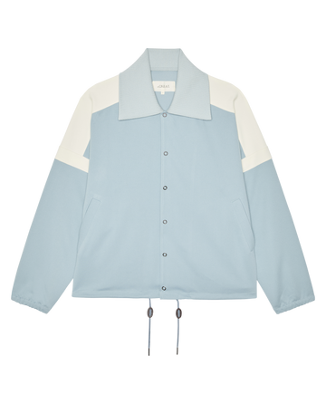 The Trainer Jacket by THE GREAT. SP26 D1 in Robin Blue features snap buttons, side pockets, drawstring hem, and elastic cuffs, shown on a plain white background.