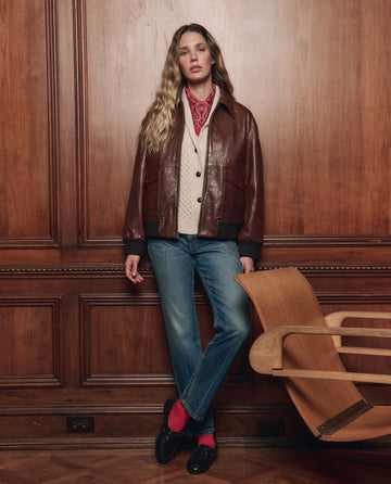 A person stands indoors against wooden paneling, wearing THE GREAT. F26 COLLECTORS The Flight Jacket in Mahogany Brown over a cream sweater, paired with blue jeans, red socks, black shoes, and a red bandana around the neck.