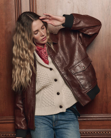 A woman with long wavy hair wears THE GREAT. F26 COLLECTORS' The Flight Jacket in Mahogany Brown, styled with a cream sweater, red bandana, and blue jeans as she poses against a wooden paneled wall.