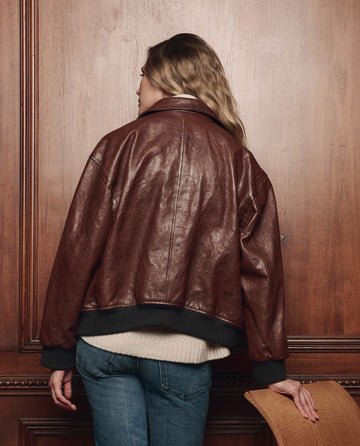Wearing THE GREAT. F26 COLLECTORS’ The Flight Jacket in Mahogany Brown, a person with wavy blonde hair and jeans stands facing a wooden wall, resting one hand on the back of a wooden chair.