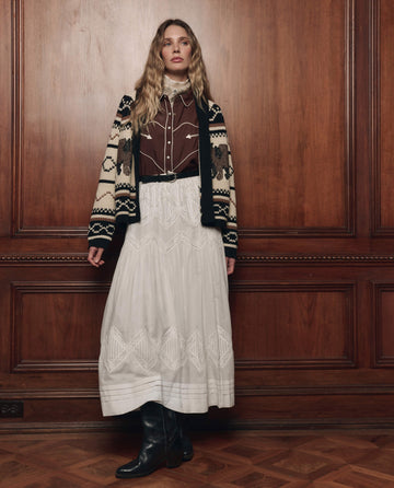 A woman stands indoors against a wood-paneled wall, wearing THE GREAT. F26 COLLECTORS The Bear Lodge Cardigan in Black, paired with a brown shirt, long white embroidered skirt, and black boots.