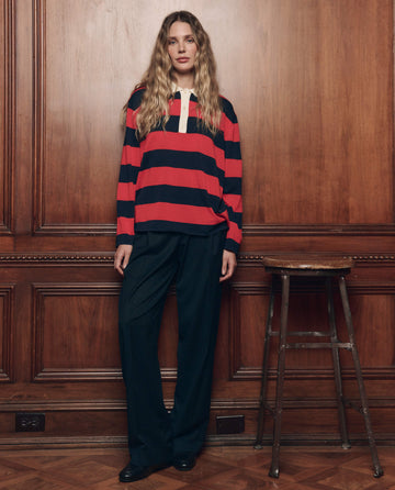 A person with long wavy hair stands indoors by a wooden wall, wearing THE GREAT. F26 COLLECTORS' The Academy Polo Sweater in Hot Red League Stripe and loose dark pants, next to a metal stool.