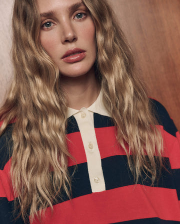 A person with long wavy blonde hair wearing THE GREAT. F26 COLLECTORS The Academy Polo Sweater in Hot Red League Stripe stands in front of a wooden background.