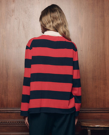 A person with long, wavy hair stands facing a wooden wall, wearing THE GREAT. F26 COLLECTORS The Academy Polo Sweater in Hot Red League Stripe and dark pants.
