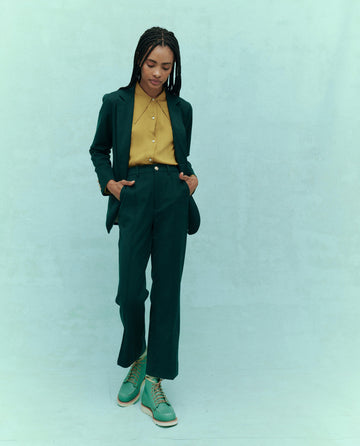 A person, embodying sophisticated fashion, dons a green suit with a yellow shirt and The Prim Trouser in Winter Pine by THE GREAT. HOL 24. Their hands are casually tucked in pockets as they stand confidently against a light green backdrop.