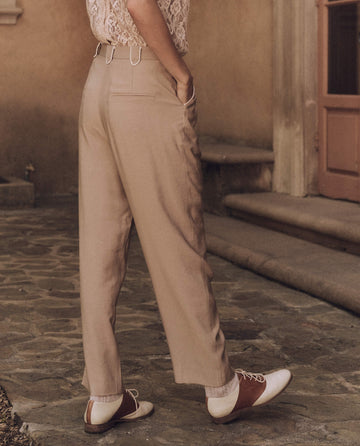 A person stands on a stone path wearing The Western Maker Trouser in Barley by THE GREAT. SP25 D1 and a lace top, paired with stylish brown and white shoes near a building entrance.
