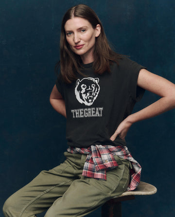 A woman sits on a stool wearing THE GREAT. SP25 COLLECTORS The Boxy Crew. Graphic tee in washed black with a bear graphic, olive pants, and a plaid shirt tied around her waist.