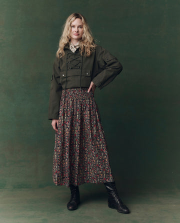 Against a green backdrop, a person wears THE GREAT. F25 D2's The Utility Pullover in Dusty Moss, paired with a floral pleated skirt, lace blouse, and black lace-up boots.
