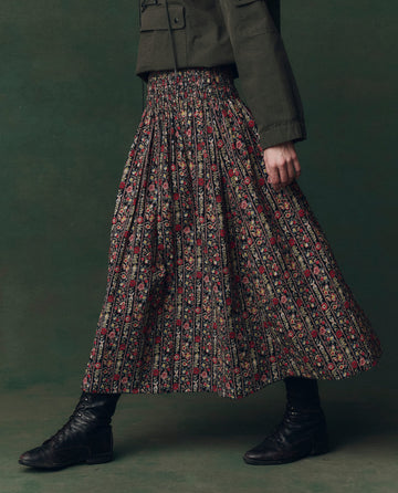 Model wears THE GREAT. F25 D2’s The Canary Skirt in Garland Print with a green jacket and black lace-up boots, walking against a dark green background.