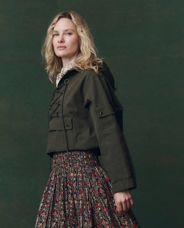 A person with wavy blonde hair wears THE GREAT. F25 D2’s The Utility Pullover in Dusty Moss, paired with a pleated floral skirt, standing against a dark green background.