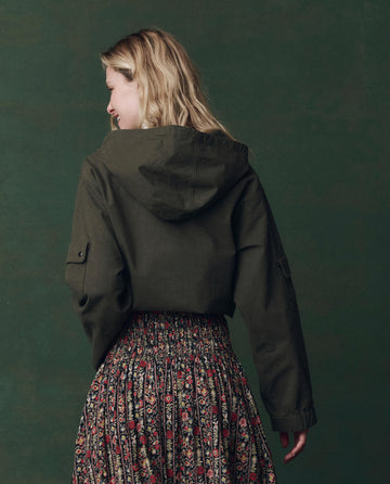 A person with long blonde hair, facing away against a dark green background, wears THE GREAT. F25 D2 The Utility Pullover in Dusty Moss and a floral-patterned skirt.