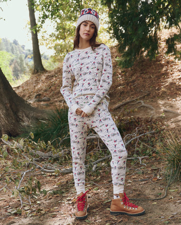 A person stands outdoors among trees and fallen leaves, wearing THE GREAT. F25 TGO The Classic Crew Thermal in Slopeside Print, paired with red hiking boots and a festive knit hat.