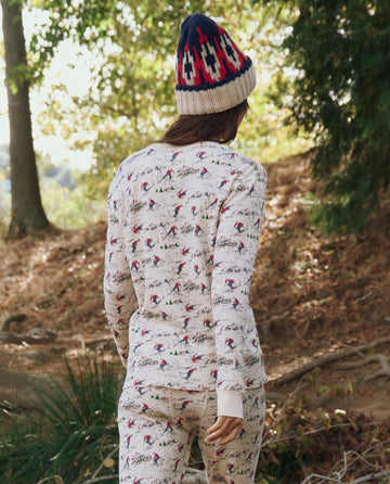 Wearing THE GREAT. F25 TGO's The Classic Crew Thermal in Slopeside Print, a person walks outdoors on a dirt path amid trees and foliage.