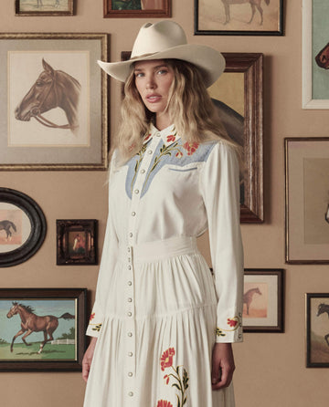 A woman in THE GREAT. STETSON SP26 The Embroidered Western Shirt—Cream w/ Chambray Mix Multi Emb—styled as a dress, stands before a beige wall adorned with framed horse paintings.
