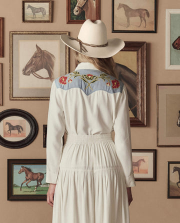 Wearing THE GREAT. STETSON SP26 The Embroidered Western Shirt in Cream W/ Chambray Mix Multi Emb, a person in a hat stands facing a wall adorned with framed horse paintings and drawings.