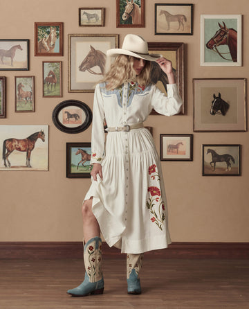 A woman stands indoors before framed horse paintings, wearing a white western dress cinched with THE GREAT. STETSON SP26 The Western Narrow Belt in Cream With Silver, along with cowboy boots and a hat.