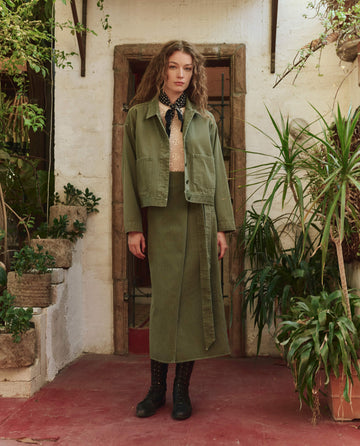 A woman stands before a rustic doorway wearing THE GREAT. PF25 The Rancher Jacket – Classic Army, styled with a skirt, beige blouse, and black scarf, surrounded by potted plants.