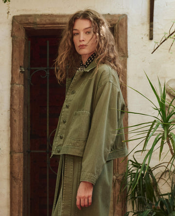 A person with curly hair stands indoors, wearing THE GREAT. PF25 The Rancher Jacket -- Classic Army and a matching skirt, in front of a stone doorway and plants.