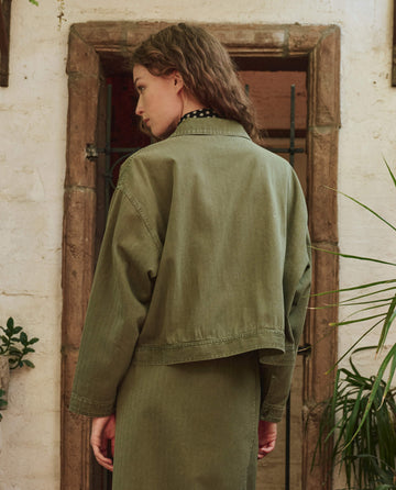 A person with wavy brown hair, seen from behind, wears THE GREAT. PF25's The Rancher Jacket – Classic Army in front of a rustic doorway framed by plants.