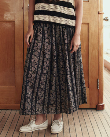 Indoors, a person wears a sleeveless striped top, white boat shoes, and THE GREAT. SU26 The Canary Skirt in Black And Cream Token Floral; their face is not visible.