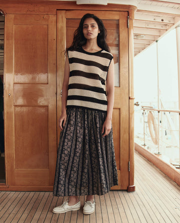 A woman stands on a wooden ship deck, facing the camera in a neutral pose. She wears THE GREAT. SU26’s The Canary Skirt in Black And Cream Token Floral, paired with a sleeveless black and white striped top and white shoes.